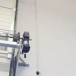 Zamontowany silnik bramy garażowej segmentowej z widoczną linką, elementami mocującymi i przewodami elektrycznymi oraz kamera przemysłowa na ścianie wewnątrz budynku.
