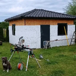 Wznoszenie małego domu z płyt warstwowych na trawniku; widoczna piła stołowa, drabina, poziomica i pies rasy pitbull.