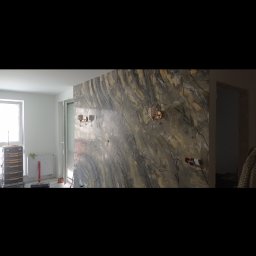 Firma Budowlana Przemysław Żurko - Ściana w pokoju z dekoracyjną, marmurkową fakturą, oświetlona przez dwa ozdobne kinkiety, widoczne gniazdka elektryczne i narzędzia budowlane w tle.