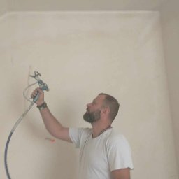 Firma Budowlana Przemysław Żurko - Pracownik w Zielonej Górze aplikuje gładź natryskową na ścianę w pomieszczeniu za pomocą pistoletu hydrodynamicznego, widoczny w trakcie pracy, z narzędziami przy pasie.