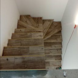 Firma Budowlana Przemysław Żurko - Widok z góry na wyłożone płytkami imitującymi drewno schody w trakcie prac wykończeniowych, z widocznymi butami i ubraniem wykonawcy.