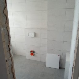 Firma Budowlana Przemysław Żurko - Wnętrze łazienki w trakcie remontu: ułożone jasne płytki ścienne z delikatnym wzorem, szare płytki podłogowe, widoczne instalacje wodno-kanalizacyjne przed montażem armatury, surowy otwór drzwiowy...