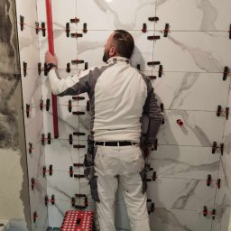 Firma Budowlana Przemysław Żurko - Mężczyzna w ubraniu roboczym, z tyłu, podczas układania płytek imitujących marmur w łazience, używając poziomicy i systemu poziomowania płytek.