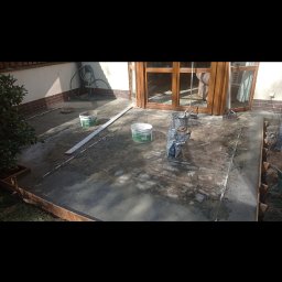 Firma Budowlana Przemysław Żurko - Przygotowanie podłoża pod taras: wylany beton, widoczne listwy poziomujące, wiaderka z zaprawą oraz worek z materiałem budowlanym przy drewnianych drzwiach tarasowych.