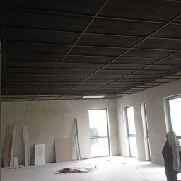 Firma Budowlana Przemysław Żurko - Wnętrze w trakcie prac wykończeniowych: surowa ściana, częściowo zamontowany sufit podwieszany z widoczną konstrukcją, narzędzia i materiały budowlane rozłożone na podłodze.