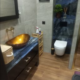 Firma Budowlana Przemysław Żurko - Nowoczesna łazienka z umywalką w kolorze bursztynu, szafką z ciemnego marmuru i drewnopodobnymi płytkami podłogowymi. Na ścianie wisi obrazek kota czytającego gazetę na toalecie.