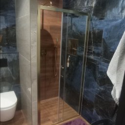 Firma Budowlana Przemysław Żurko - Wykończona łazienka w Zielonej Górze z kabiną prysznicową ze złotymi ramami, marmurowymi płytkami w odcieniach granatu i brązu oraz drewnopodobną podłogą.