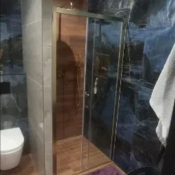 Wykończona łazienka w Zielonej Górze z kabiną prysznicową ze złotymi ramami, marmurowymi płytkami w odcieniach granatu i brązu oraz drewnopodobną podłogą.