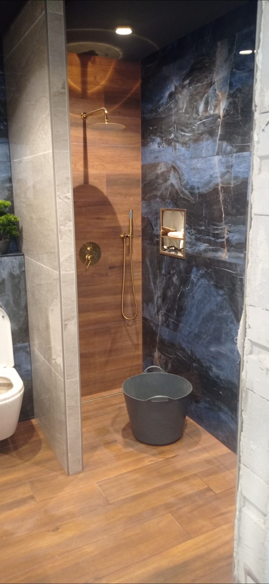 Nowoczesna kabina prysznicowa z panelami imitującymi drewno i marmur, złota armatura, widoczne wiadro na podłodze i fragment toalety, wnętrze w trakcie wykańczania.