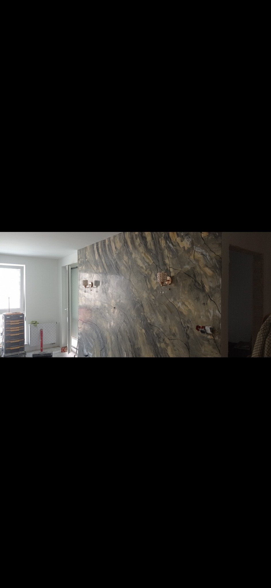 Ściana w pokoju z dekoracyjną, marmurkową fakturą, oświetlona przez dwa ozdobne kinkiety, widoczne gniazdka elektryczne i narzędzia budowlane w tle.