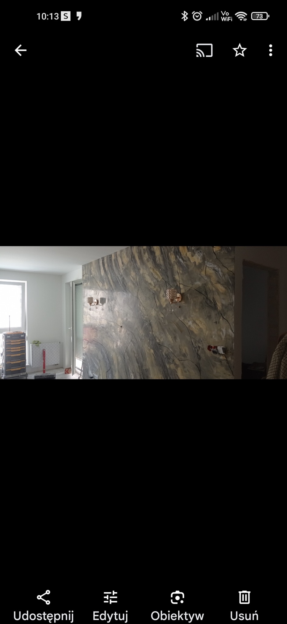 Ściana z efektem dekoracyjnym imitującym marmur, z zamontowanymi kinkietami, widoczne przygotowania do instalacji elektrycznej, w tle okno i grzejnik.