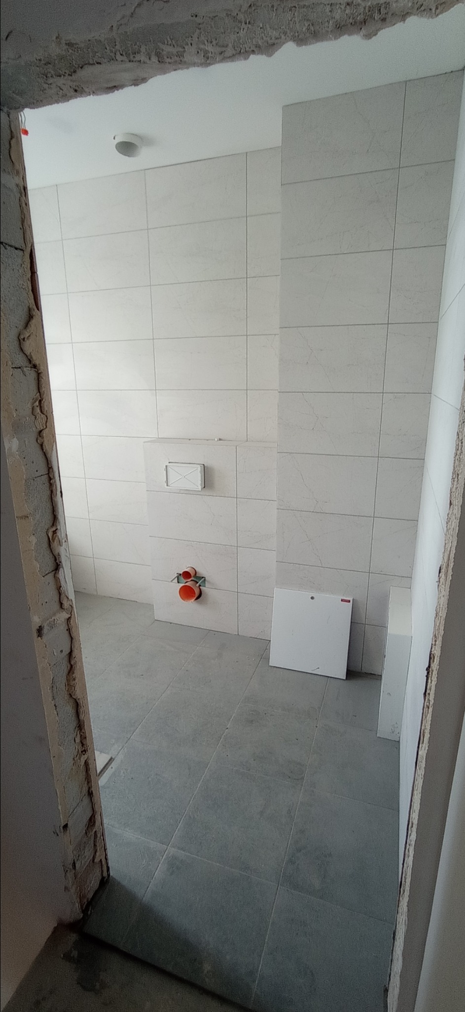 Wnętrze łazienki w trakcie remontu: ułożone jasne płytki ścienne z delikatnym wzorem, szare płytki podłogowe, widoczne instalacje wodno-kanalizacyjne przed montażem armatury, surowy otwór drzwiowy...