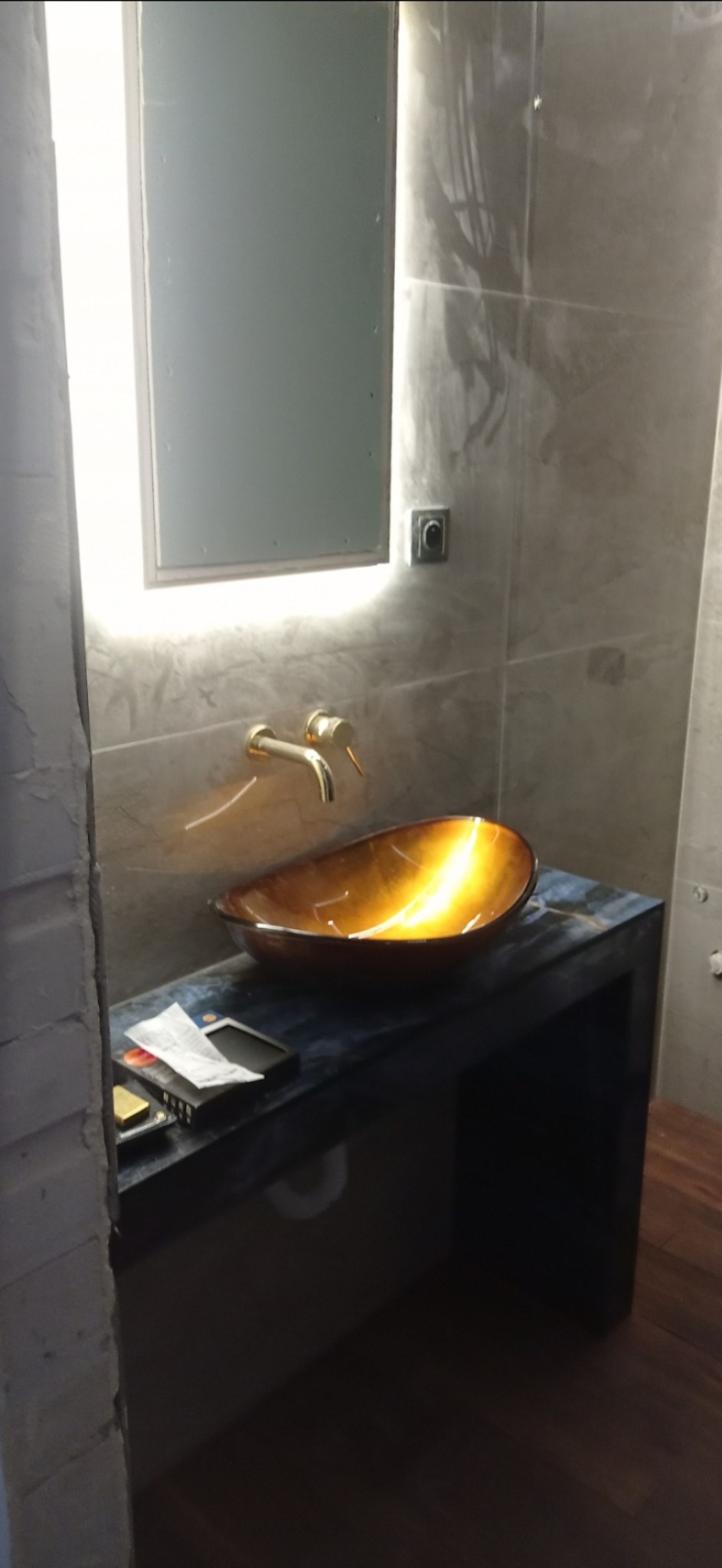 Umywalka w odcieniach bursztynu, złota bateria, podświetlane lustro i ściany wykończone w szarościach, widoczne materiały budowlane na blacie.