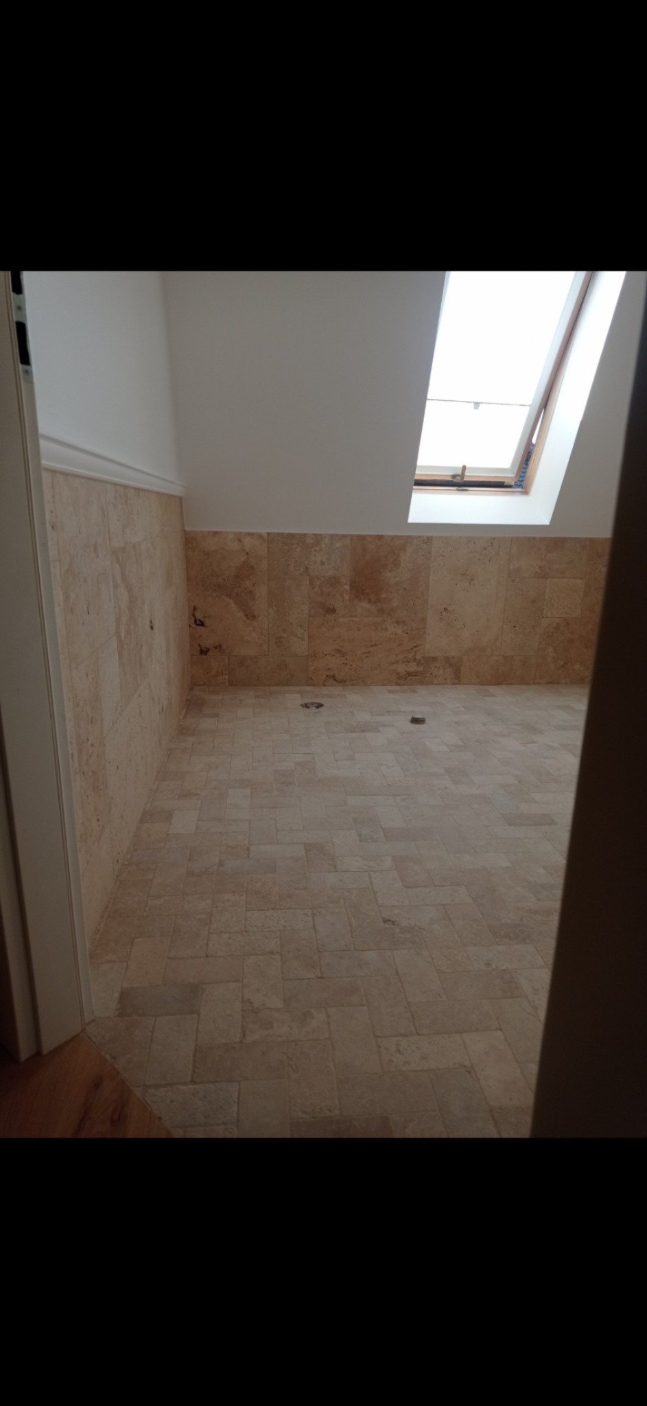 Wykończona łazienka na poddaszu z beżowymi płytkami na podłodze i ścianach do połowy wysokości, widoczne dwa odpływy w podłodze, okno dachowe w skosie.