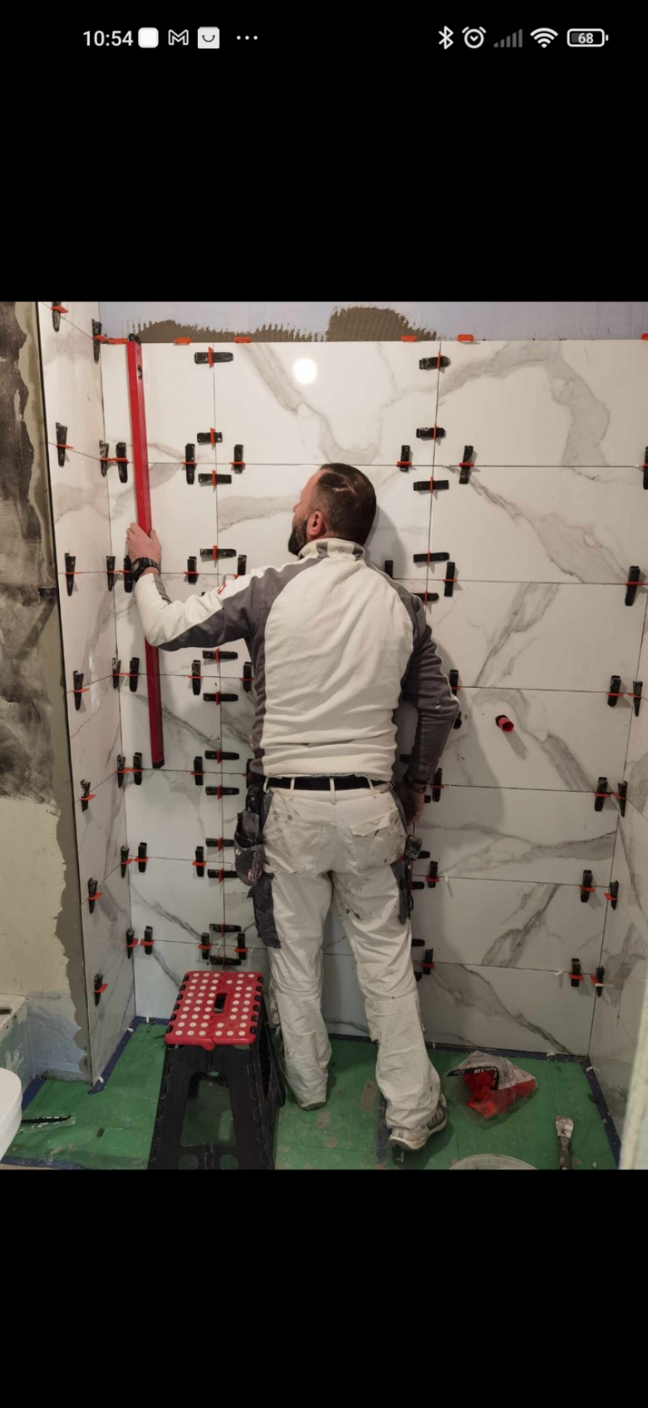 Mężczyzna w ubraniu roboczym, z tyłu, podczas układania płytek imitujących marmur w łazience, używając poziomicy i systemu poziomowania płytek.