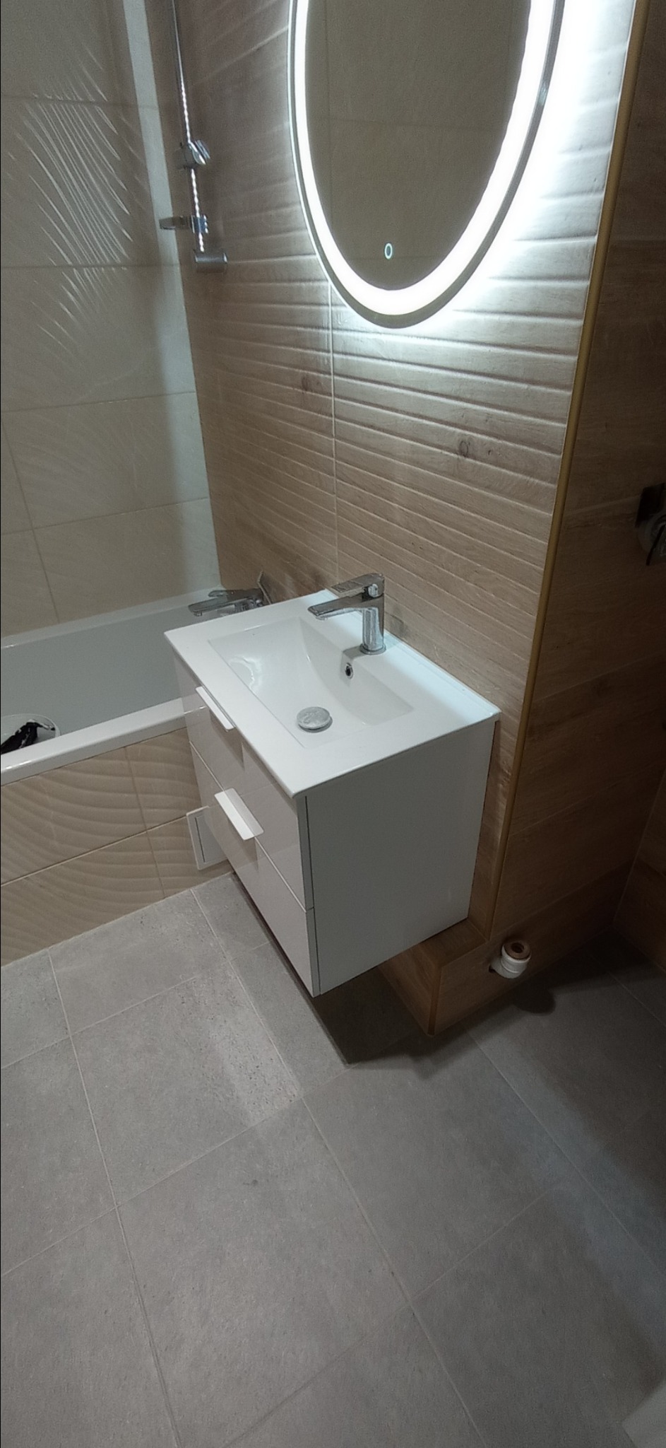 Nowoczesna łazienka z białą szafką pod umywalkę, podświetlanym okrągłym lustrem i beżowymi płytkami imitującymi drewno na ścianach.