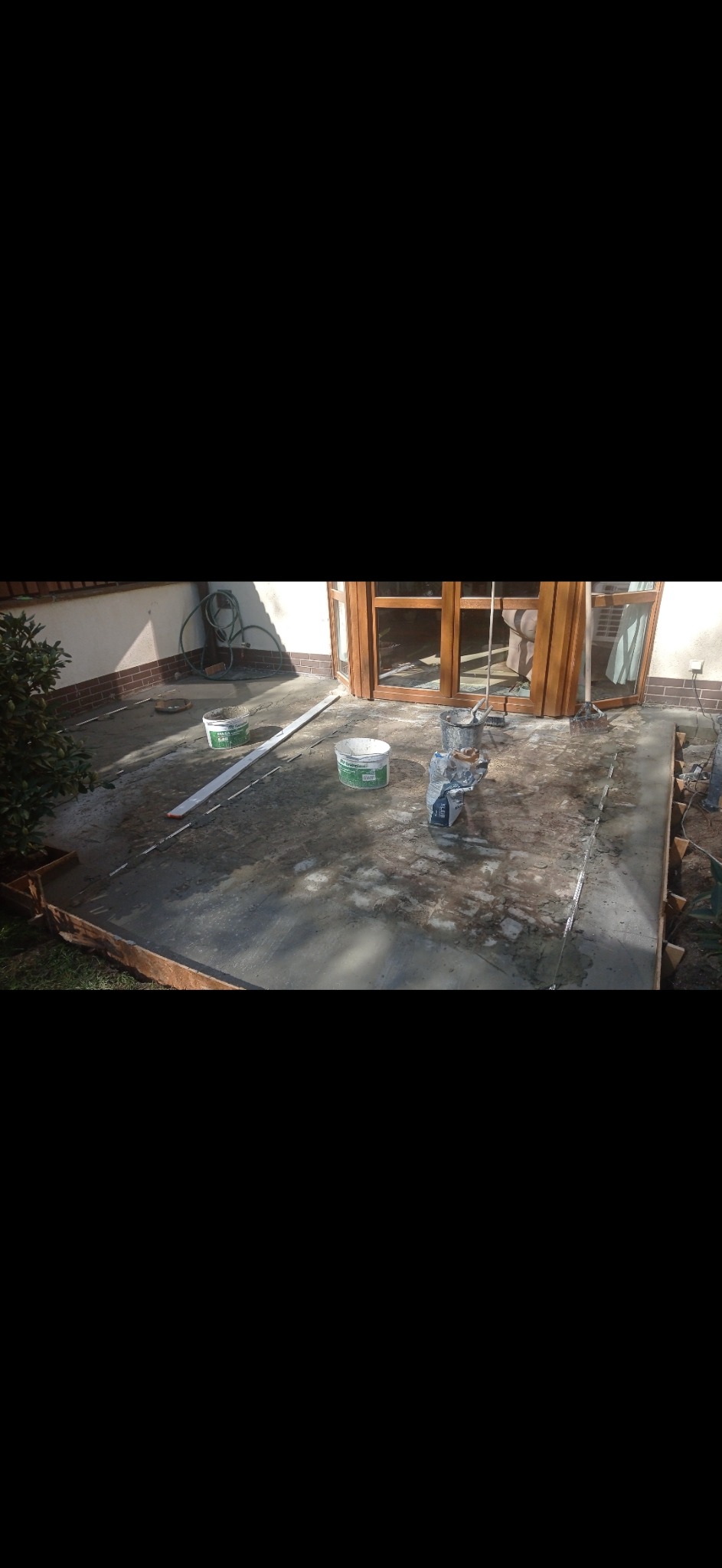 Przygotowanie podłoża pod taras: wylany beton, widoczne listwy poziomujące, wiaderka z zaprawą oraz worek z materiałem budowlanym przy drewnianych drzwiach tarasowych.