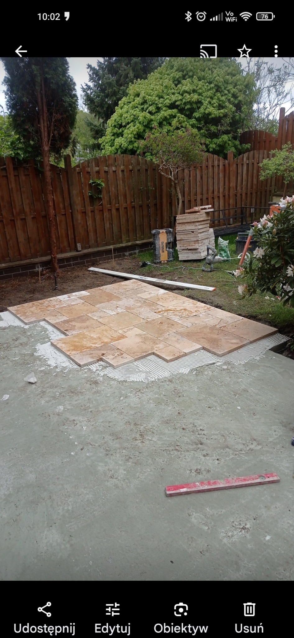 Układanie trawertynu na tarasie w ogrodzie, widoczny klej i poziomica, w tle drewniany płot i zieleń.