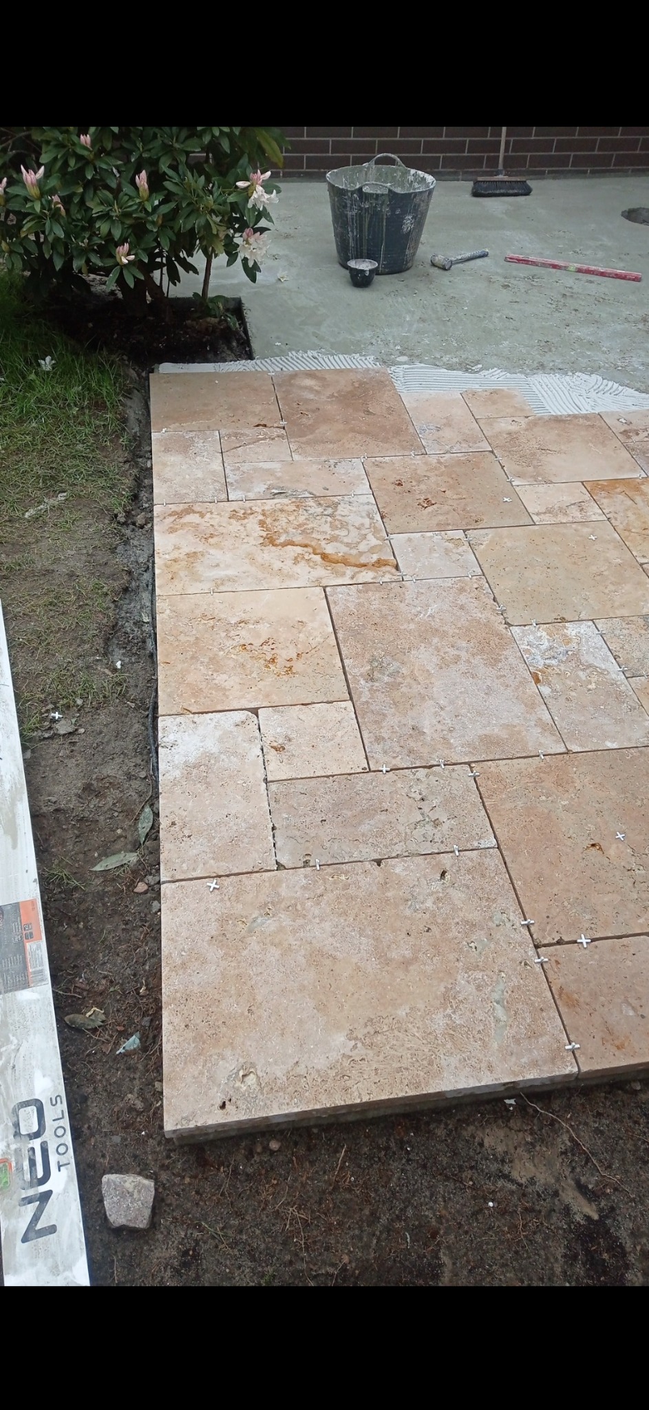 Układanie trawertynu w ogrodzie, widoczne krzyżyki dystansowe, wiadro z zaprawą oraz narzędzia brukarskie w tle