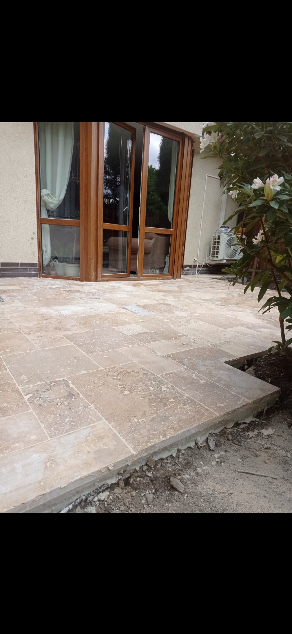 Wyłożony trawertynem taras z otwartymi, drewnianymi drzwiami balkonowymi i widocznym klimatyzatorem na ścianie budynku.