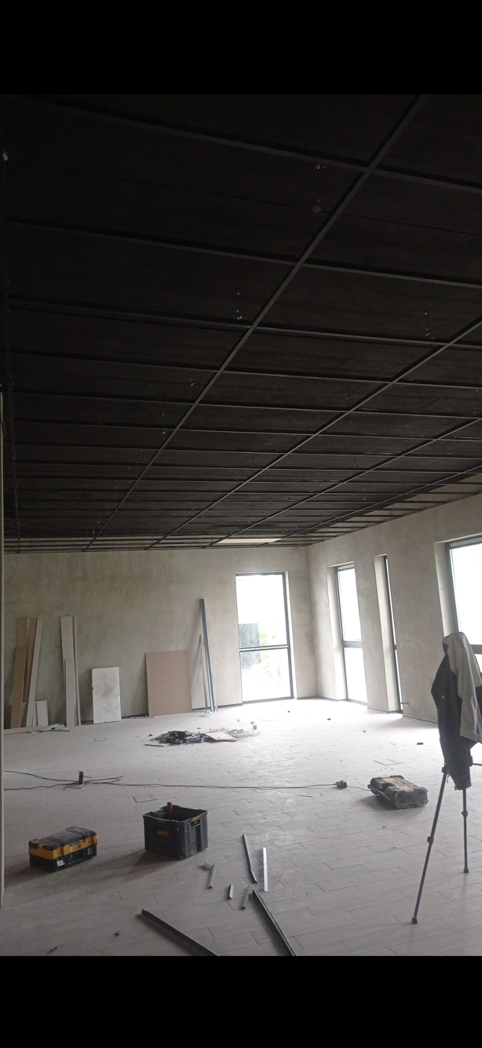 Wnętrze w trakcie prac wykończeniowych: surowa ściana, częściowo zamontowany sufit podwieszany z widoczną konstrukcją, narzędzia i materiały budowlane rozłożone na podłodze.