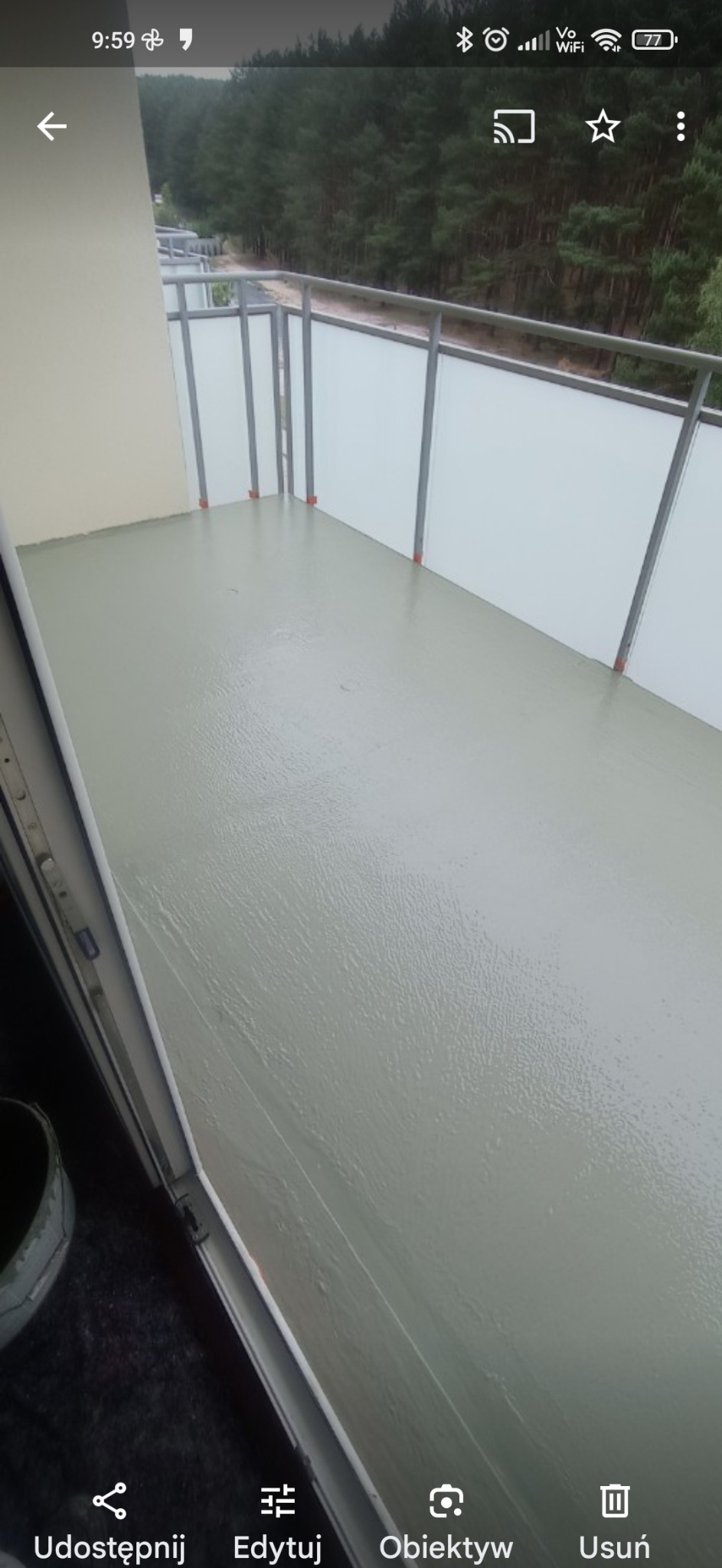 Balkon po wylaniu samopoziomującej wylewki, widoczne mokre podłoże i szara balustrada z białym wypełnieniem, w tle las.