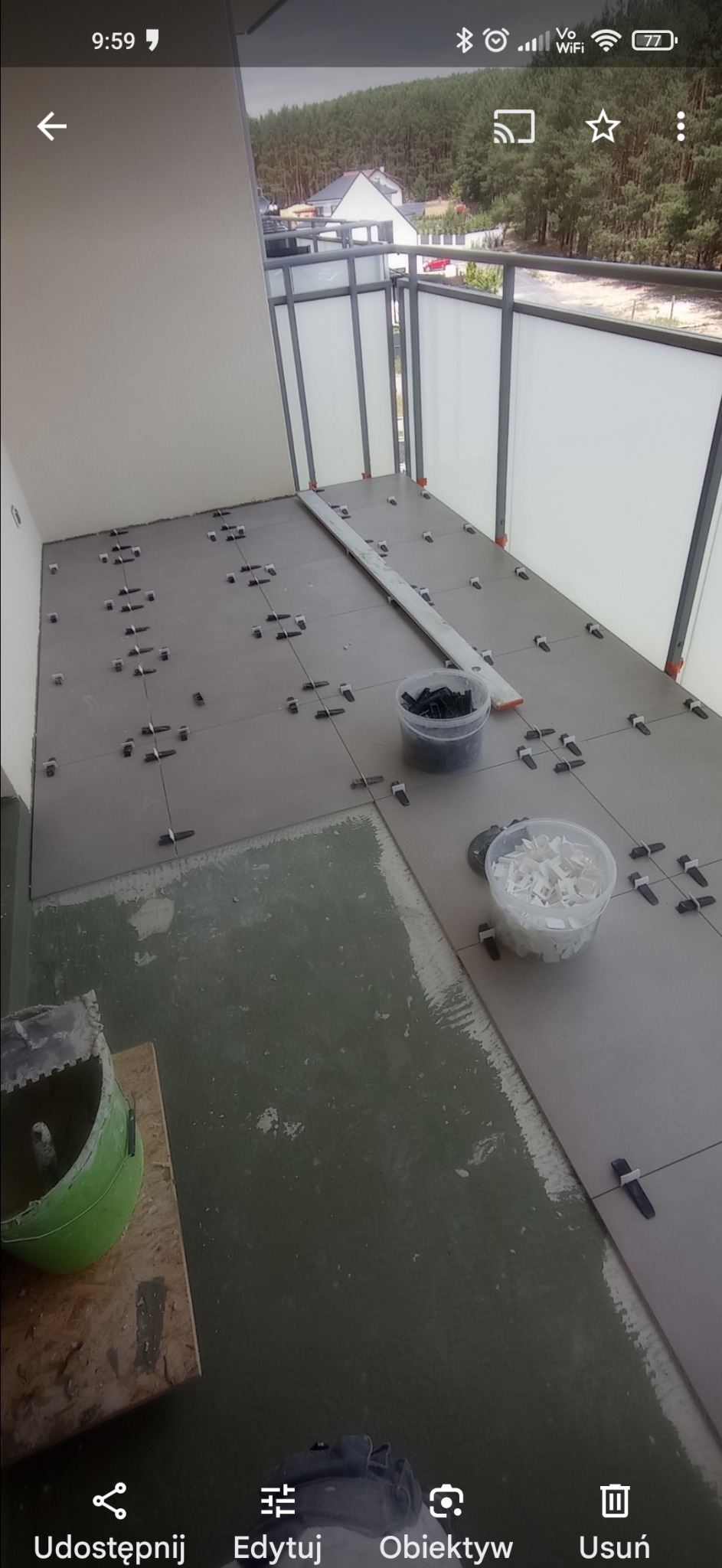 Układanie szarych płytek na balkonie z użyciem krzyżyków dystansowych, widoczny poziomica i pojemniki z krzyżykami, w tle balustrada i zieleń.