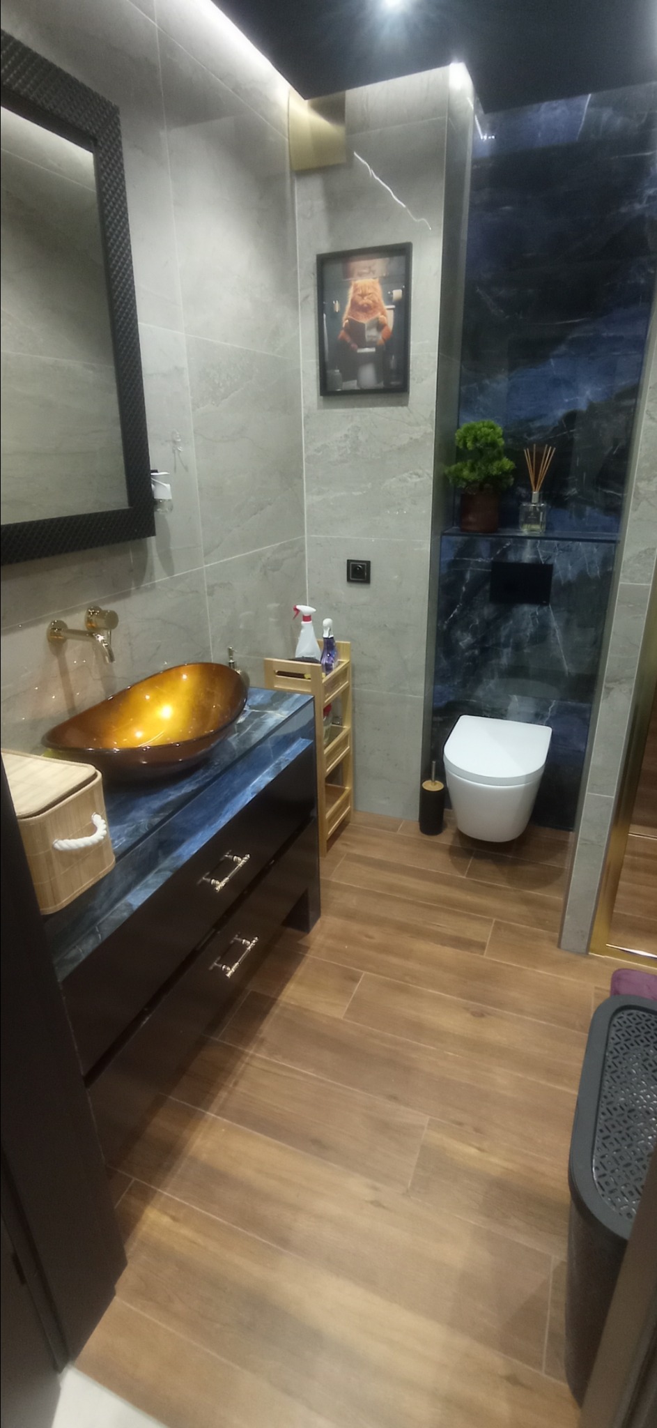 Nowoczesna łazienka z umywalką w kolorze bursztynu, szafką z ciemnego marmuru i drewnopodobnymi płytkami podłogowymi. Na ścianie wisi obrazek kota czytającego gazetę na toalecie.