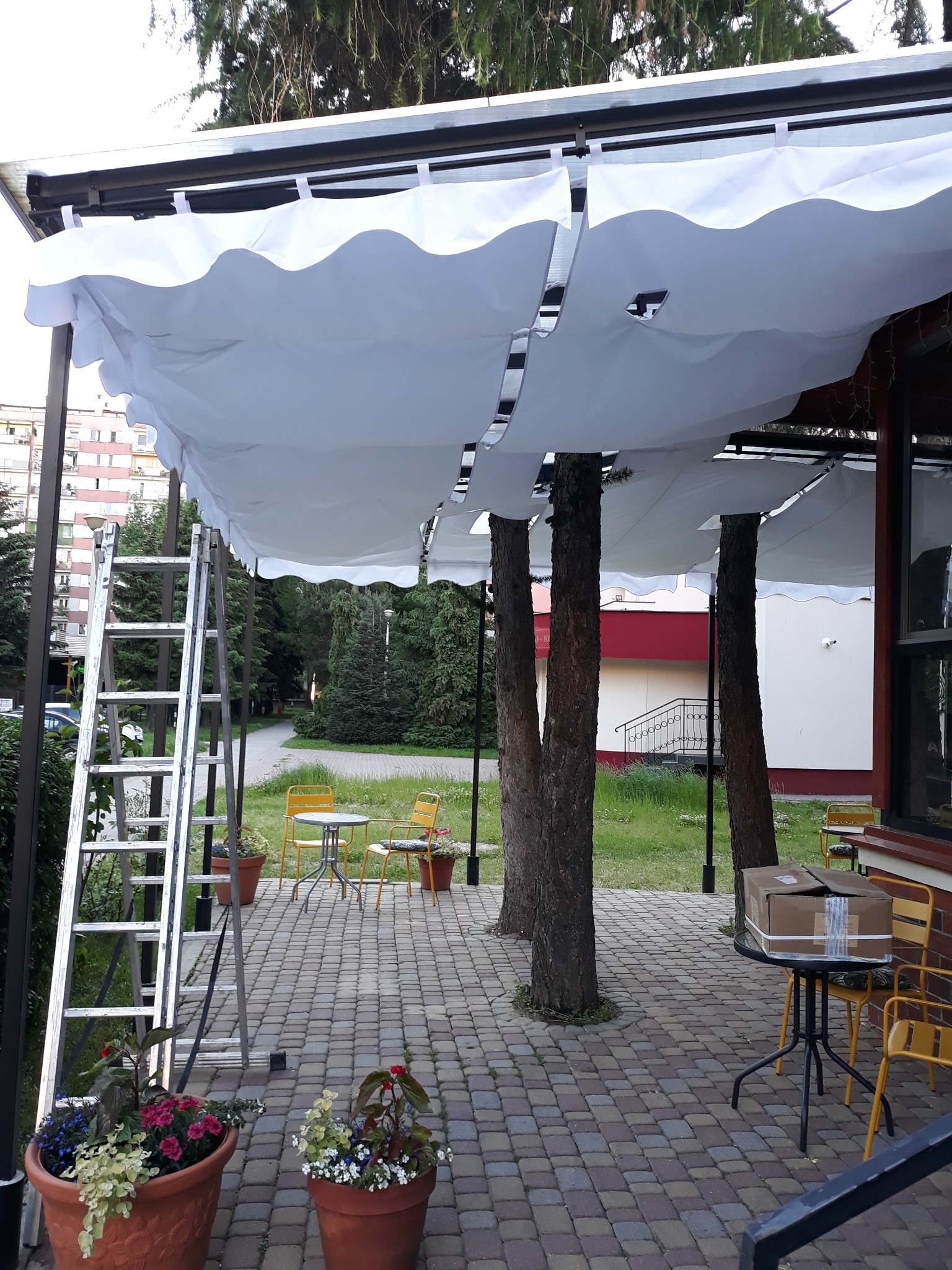 Montaż białego, falistego zadaszenia tarasu z otworami na pnie drzew, drabina oparta o konstrukcję, stolik z kartonami, żółte krzesła, donice z kwiatami, kostka brukowa.