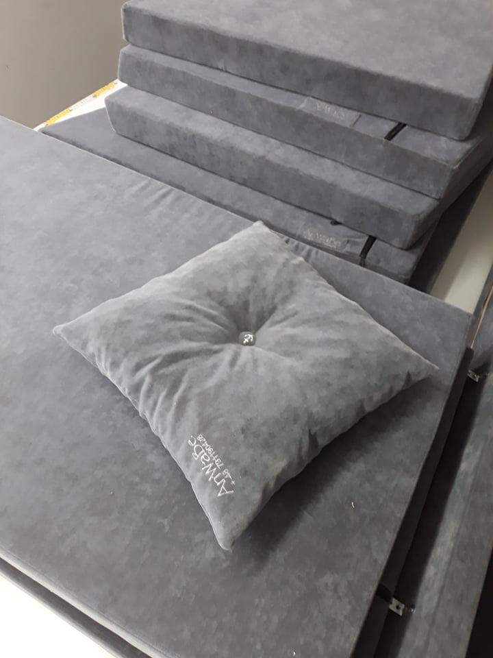 Stos szarych, welurowych poduszek i materacy z widocznym logo firmy na jednej z poduszek, częściowo złożone na stole.