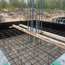 Zbrojenie fundamentów z wystającymi prętami pionowymi, szalunek z czarnej płyty, drewniane podpory, widok z góry na siatkę zbrojeniową.