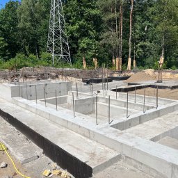 2M EDIFICE - Wylane fundamenty domu jednorodzinnego z wystającymi prętami zbrojeniowymi, w tle las i słup wysokiego napięcia.