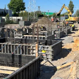 Budowa fundamentów: Wykop z szalunkami i zbrojeniem, w tle koparka Komatsu i pracownicy w kaskach.