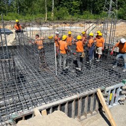 2M EDIFICE - Grupa robotników w pomarańczowych koszulkach i kaskach montuje zbrojenie fundamentu pod budowę, widoczne szalunki i siatka z prętów stalowych, w tle drzewa i linia wysokiego napięcia.