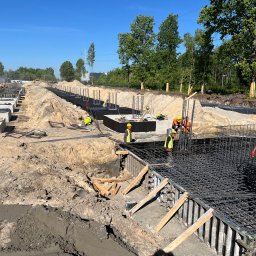 2M EDIFICE - Prace zbrojarskie na budowie fundamentów: widok na zbrojenie w szalunkach, pracownicy w odblaskowych kamizelkach i kaskach, teren budowy z wykopami i drzewami w tle.
