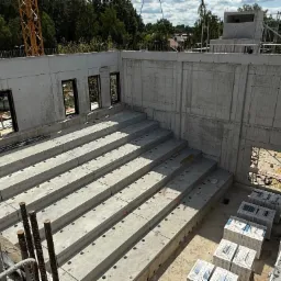 Budowa budynku żelbetowego: surowe, szare ściany z otworami okiennymi i stopniowanymi fundamentami. Żuraw budowlany w tle, widok z góry.