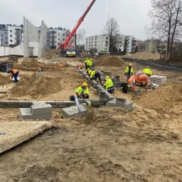 Budowa osiedla mieszkaniowego w trakcie realizacji: robotnicy w ż&oacute;łtych kamizelkach układają bloczki betonowe na fundamencie, w tle żuraw budowlany i budynki mieszkalne.