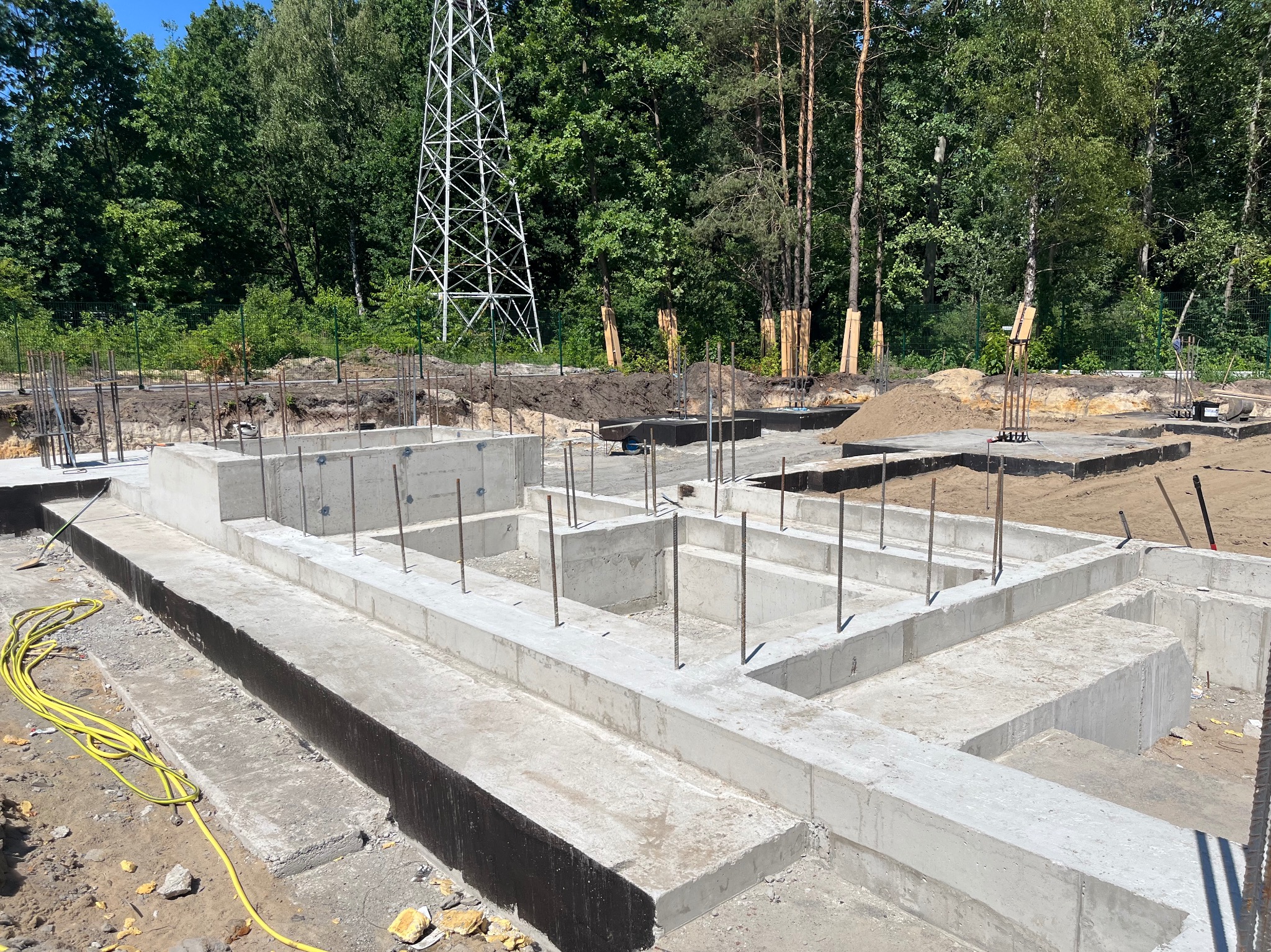 Wylane fundamenty domu jednorodzinnego z wystającymi prętami zbrojeniowymi, w tle las i słup wysokiego napięcia.