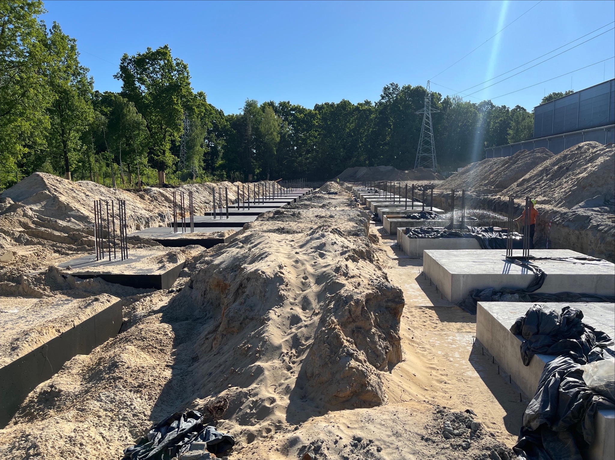 Wykop pod fundamenty z przygotowanymi zbrojeniami i izolacją, widoczny pracownik w oddali, teren budowy w słoneczny dzień.