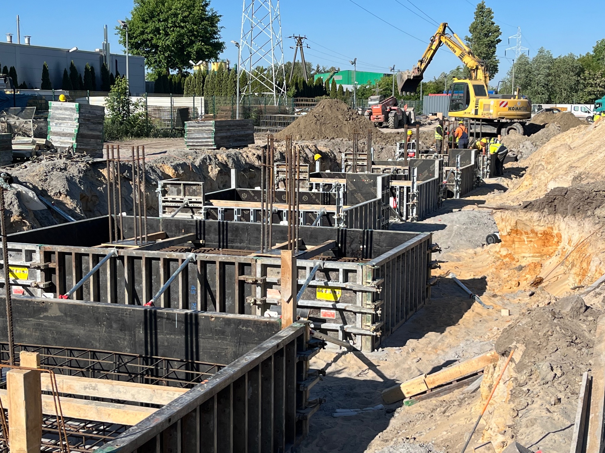 Budowa fundamentów: Wykop z szalunkami i zbrojeniem, w tle koparka Komatsu i pracownicy w kaskach.