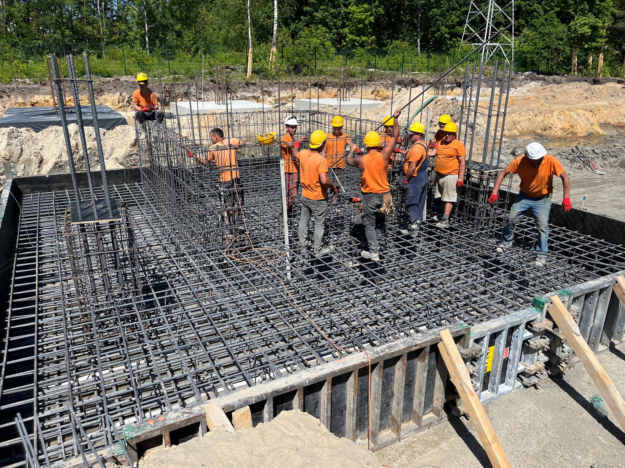 Grupa robotników w pomarańczowych koszulkach i kaskach montuje zbrojenie fundamentu pod budowę, widoczne szalunki i siatka z prętów stalowych, w tle drzewa i linia wysokiego napięcia.