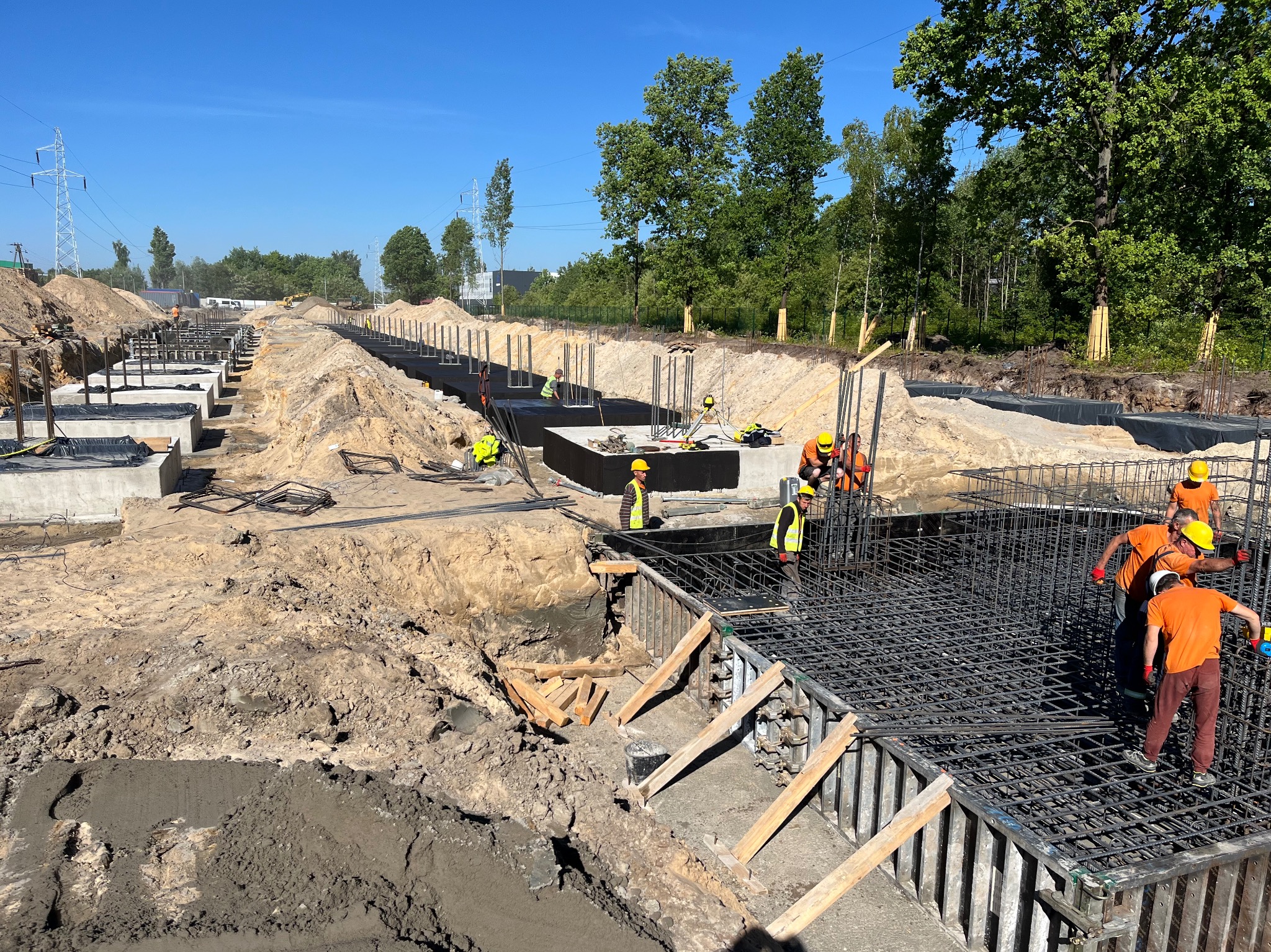 Prace zbrojarskie na budowie fundamentów: widok na zbrojenie w szalunkach, pracownicy w odblaskowych kamizelkach i kaskach, teren budowy z wykopami i drzewami w tle.