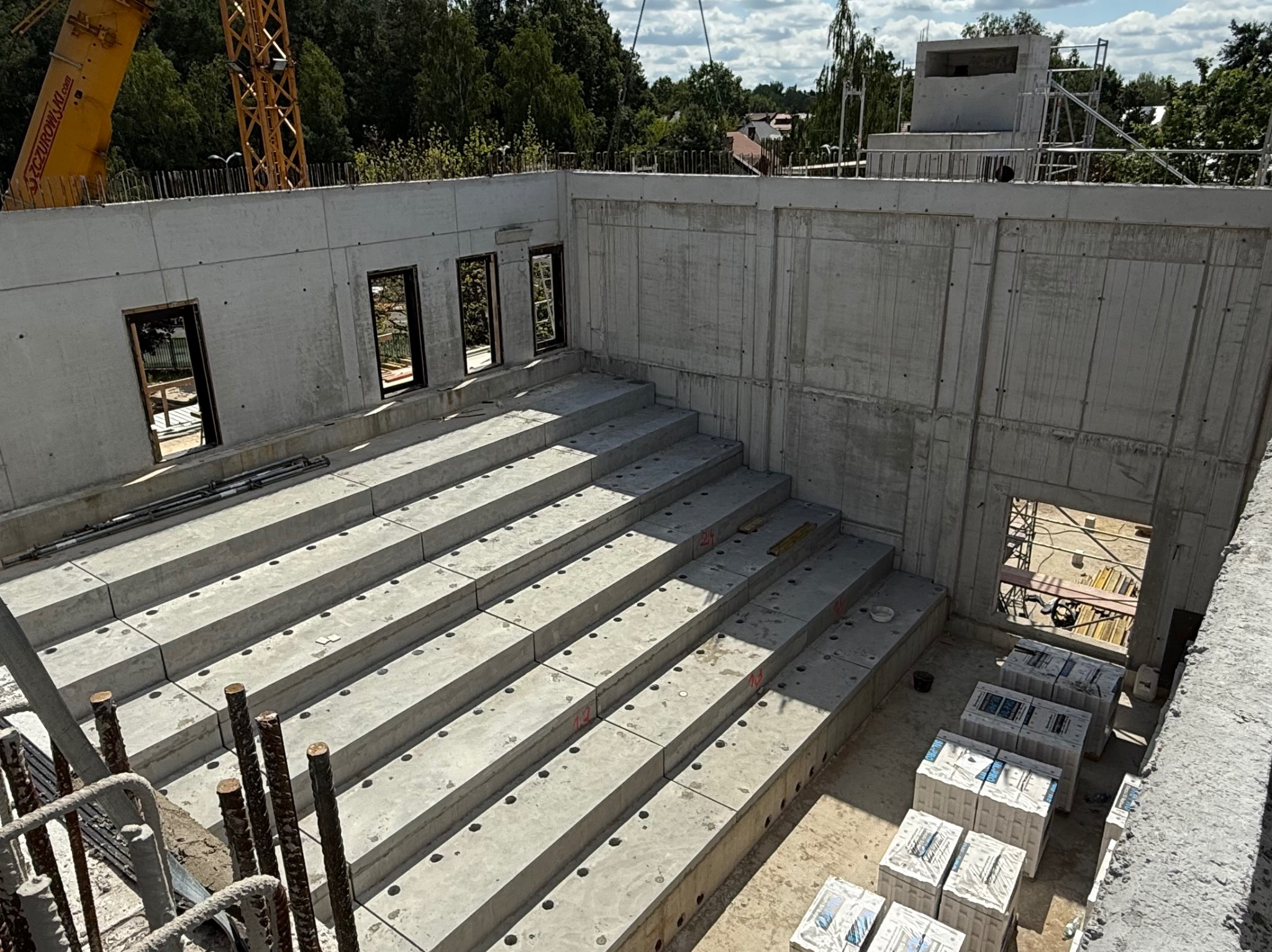 Budowa budynku żelbetowego: surowe, szare ściany z otworami okiennymi i stopniowanymi fundamentami. Żuraw budowlany w tle, widok z góry.