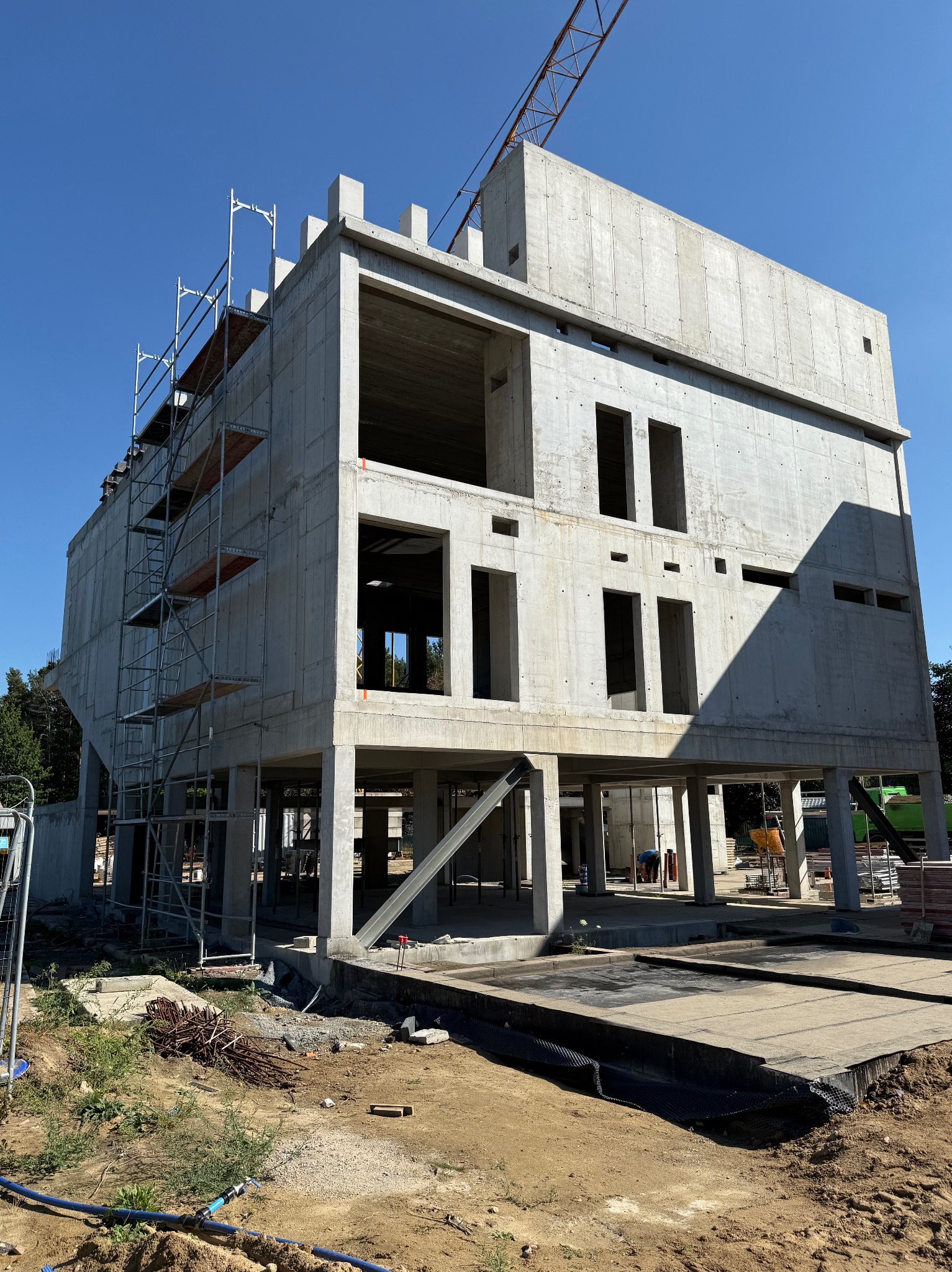 Surowy, betonowy budynek w trakcie budowy, z widocznymi rusztowaniami i żurawiem w tle, ukazujący postęp prac konstrukcyjnych w słoneczny dzień.