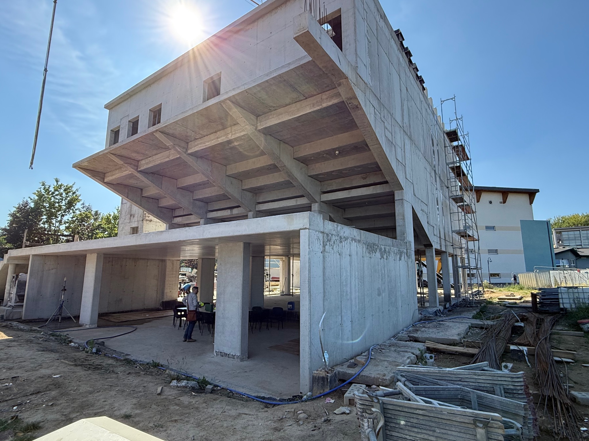 Surowy budynek żelbetowy w trakcie budowy, z widocznym szkieletem konstrukcyjnym, rusztowaniem i pracownikami. Słońce w kadrze.