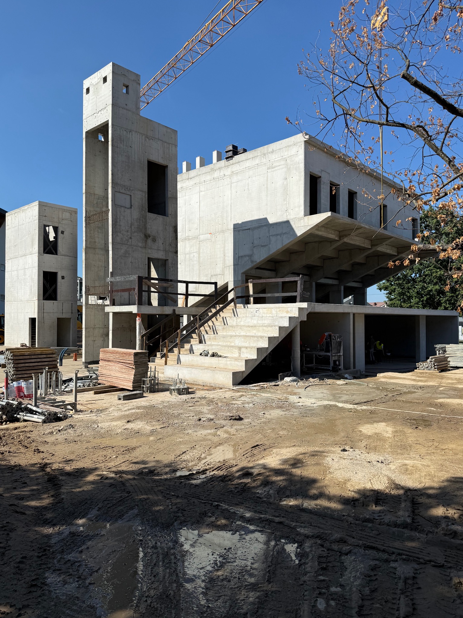 Surowy, dwupiętrowy budynek żelbetowy w trakcie budowy w słoneczny dzień. Widoczne schody zewnętrzne, szalunki, dźwig w tle i dwóch pracowników wewnątrz budynku.