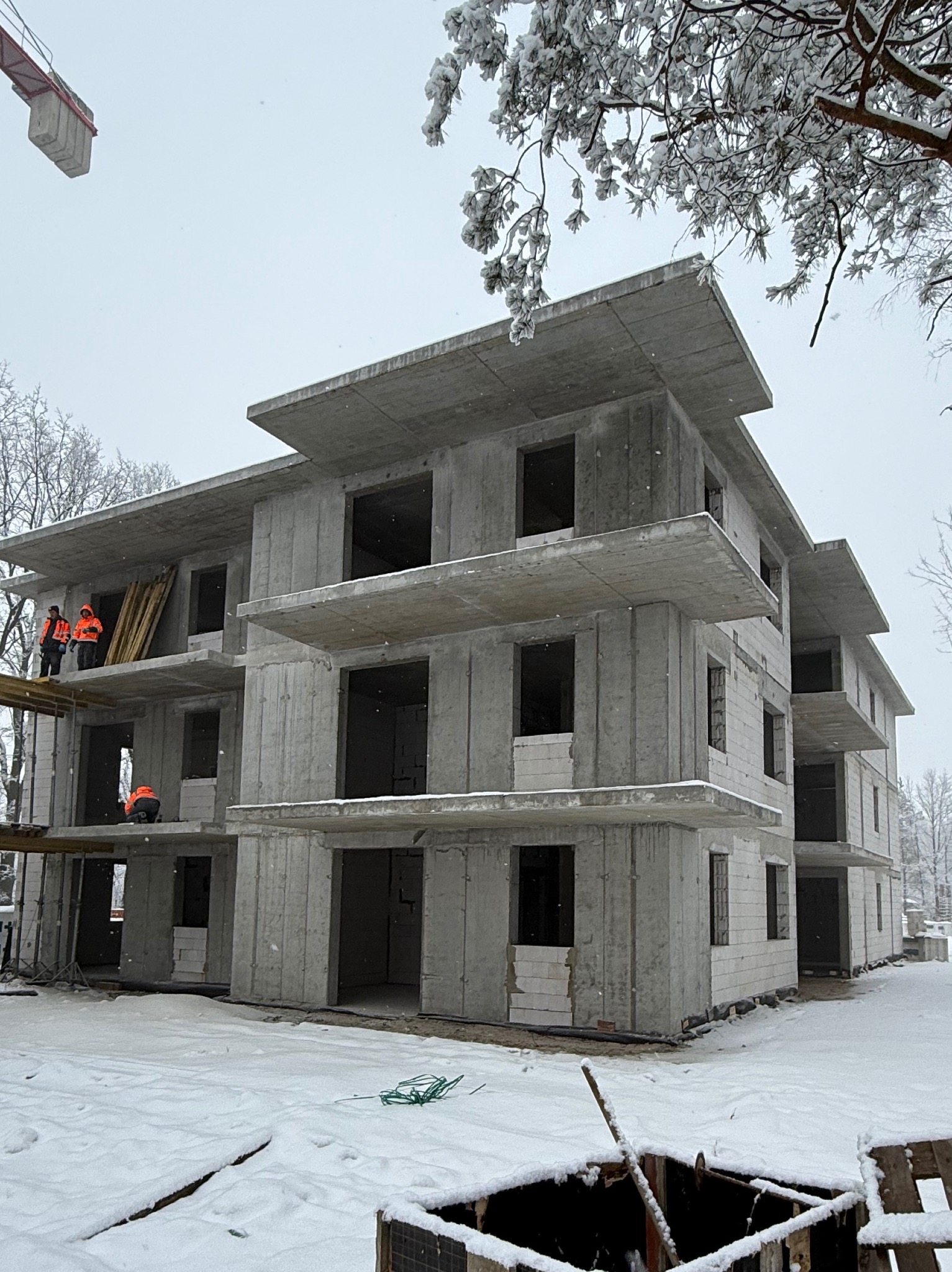 Surowy, trzypiętrowy budynek z betonu w zimowej scenerii. Widoczni robotnicy w pomarańczowych kamizelkach na jednym z pięter. Częściowo ośnieżone drzewa w tle.