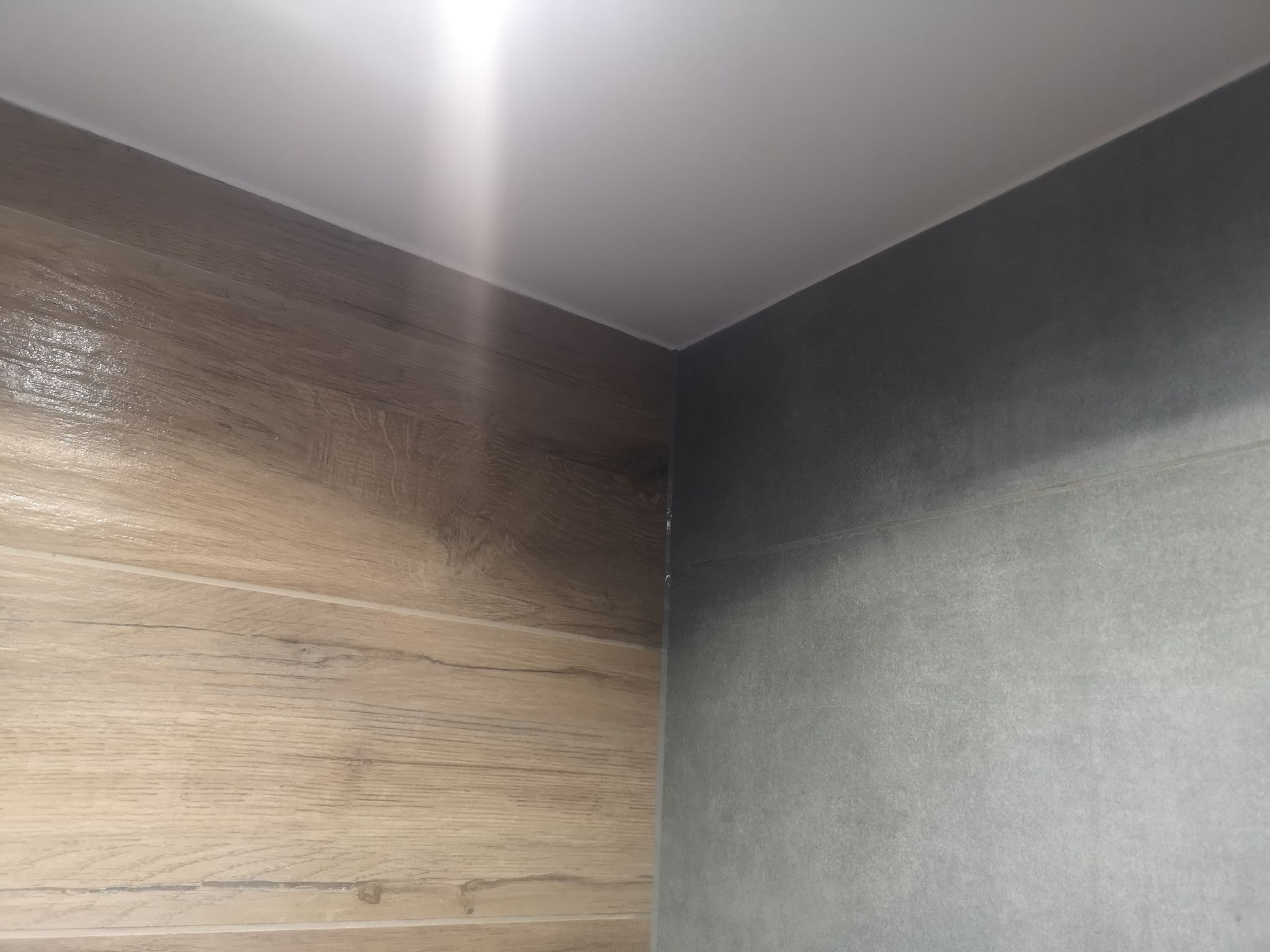 Szczegół narożnika łazienki z połączeniem płytek imitujących drewno i szarych płytek oraz białego sufitu podwieszanego.