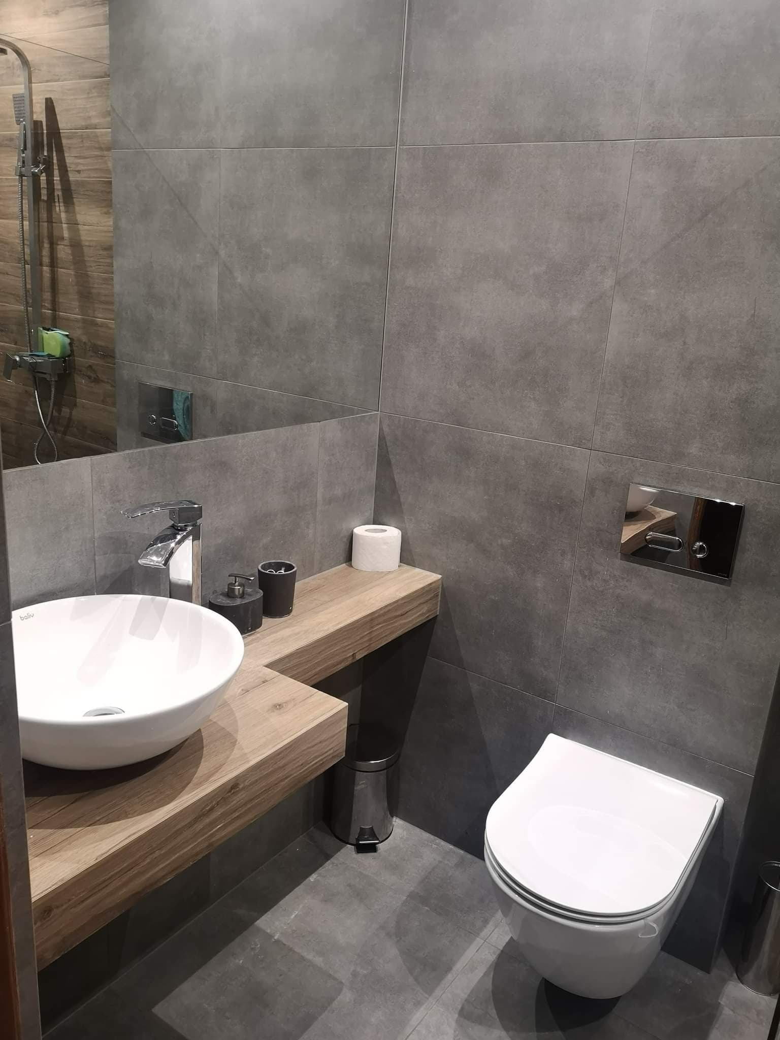 Nowoczesna łazienka z szarymi, wielkoformatowymi płytkami, wiszącą miską WC, umywalką nablatową na drewnianym blacie i chromowaną armaturą.