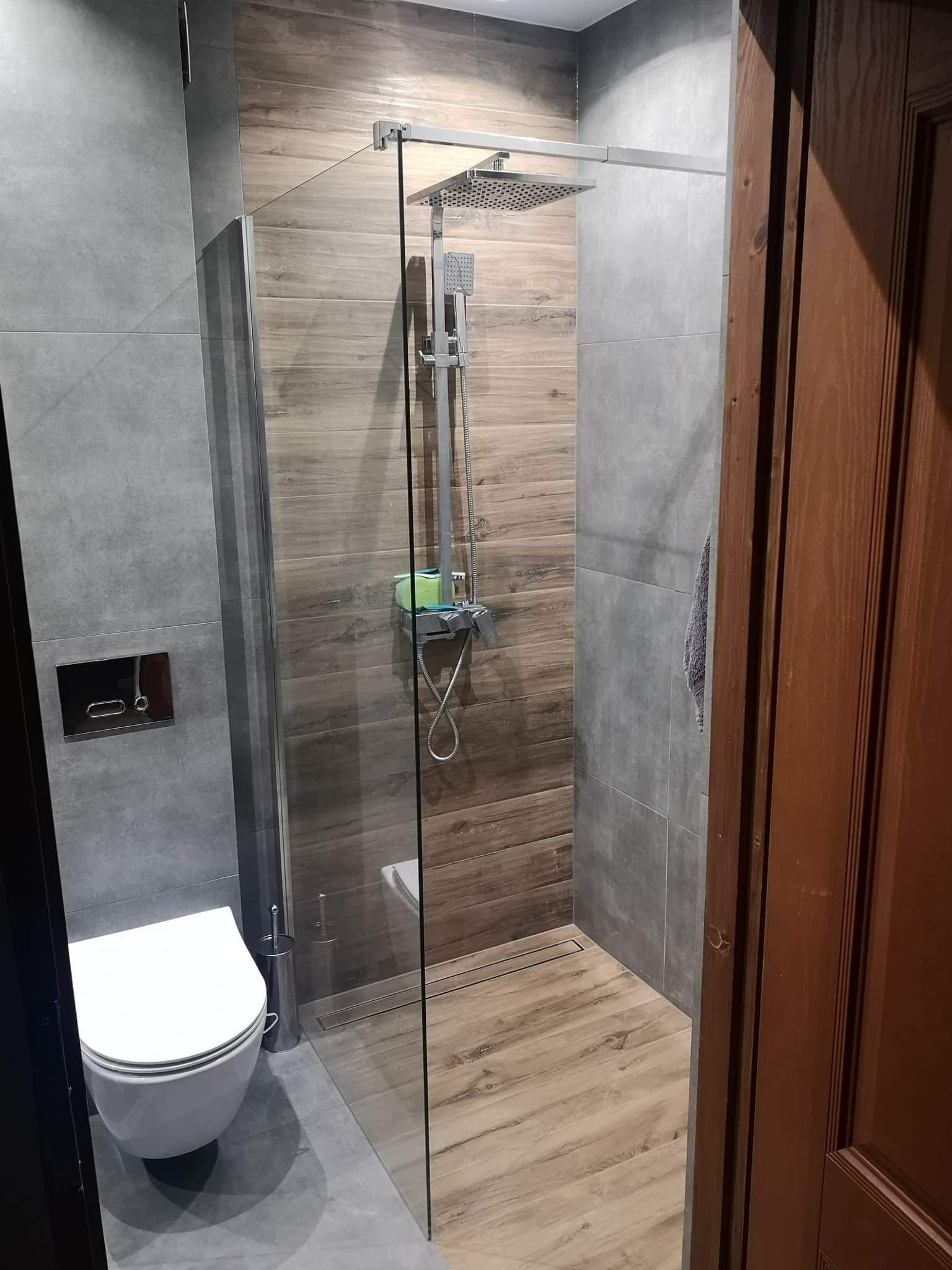 Nowoczesna łazienka z kabiną prysznicową typu walk-in, wyłożona szarymi i drewnopodobnymi płytkami, widoczna toaleta wisząca i brązowe drzwi.